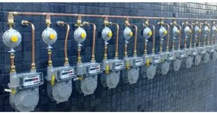 Fileira de medidores de gás com reguladores amarelos e tubulação de cobre, presos a uma parede de pastilhas azul-escuras.
